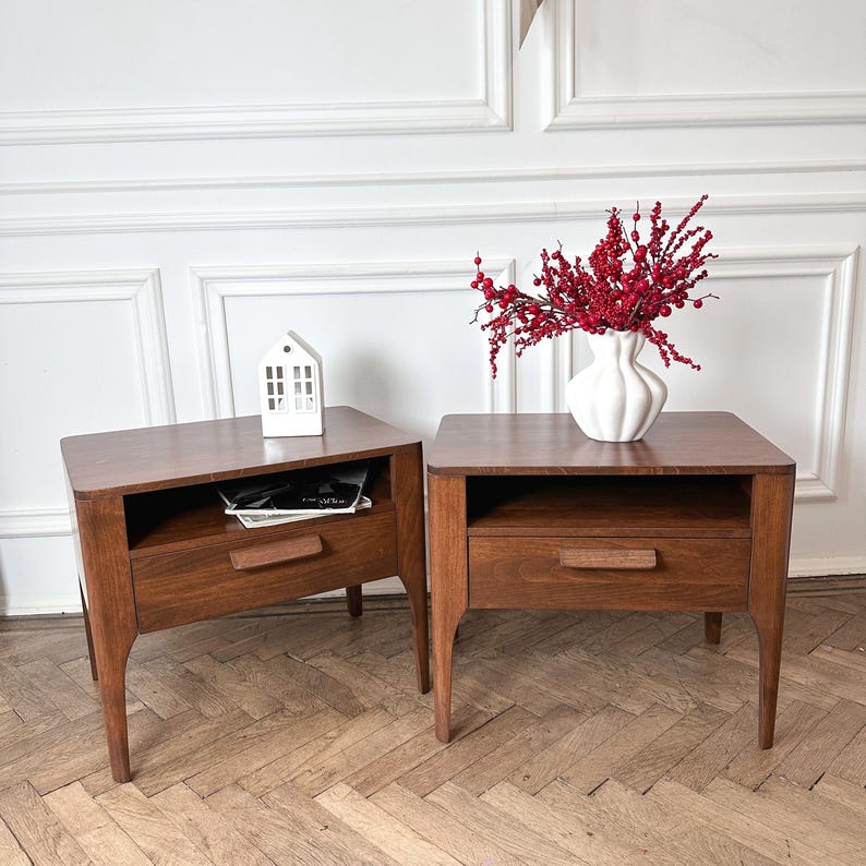 May include: Two matching dark brown wooden bedside tables, each featuring a single drawer with a horizontal handle and an open storage shelf above. A white ceramic vase filled with red berries sits on one, and a small white house model on the other.