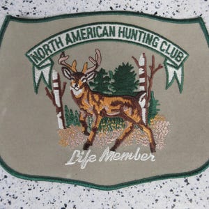 May include: Embroidered patch with a shield shape, featuring a deer with antlers, trees, and the text "NORTH AMERICAN HUNTING CLUB" and "Life Member". The patch has a green border and is primarily tan and brown.
