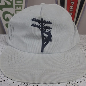 May include: A light gray corduroy baseball cap featuring a black graphic of a utility pole and a lineman. The cap has a curved brim and a structured crown. The design is centered on the front of the cap.
