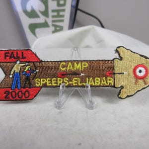 May include: An embroidered arrow-shaped patch featuring the text CAMP SPEERS-ELJABAR, FALL, and 2000. The design includes two figures using bows and arrows, a brown shaft, and a tan arrowhead pointing toward a small red and white circular target.