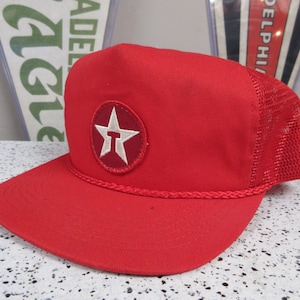 May include: Red trucker hat with a white star emblem inside a white circle. The hat has a mesh back and a curved brim. A braided rope detail accents the brim.