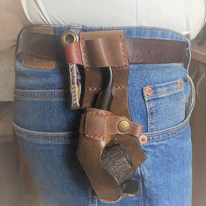 May include: A brown leather pipe holster with a metal belt loop and a brass button. The holster is attached to a brown leather belt and is worn on a person's waist. The holster is designed to hold a pipe.