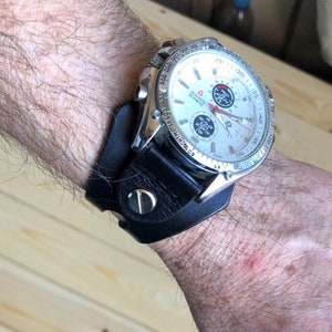 May include: A silver wristwatch with a black leather band. The watch face has a white background with black markings and a silver second hand. The watch has a chronograph feature.