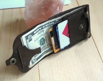 May include: A brown leather wallet with a flap that folds over to reveal a money clip and card slots. The wallet is open and shows a dollar bill and several credit cards.