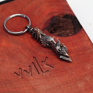 May include: A silver-toned metal keychain featuring a detailed wolf head design with a ring attached. The keychain is on a wooden surface with the word "WOLF" carved into it.