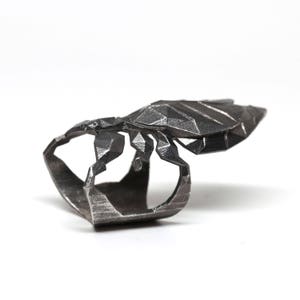 May include: A silver ring featuring a geometric scorpion design. The scorpion is detailed and has a textured finish.