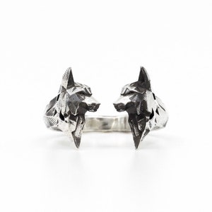 Könnte beinhalten: Ein silberfarbener Ring mit zwei stilisierten Wolfsköpfen, die einander zugewandt sind. Die Wolfsköpfe haben ein geometrisches, eckiges Design. Der Ring ist vor einem weißen Hintergrund platziert.