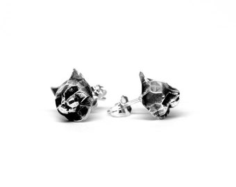 May include: A pair of silver earrings featuring a detailed, textured, abstract design. The earrings are in the shape of a head with a pointed top.