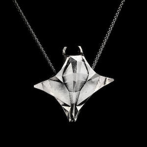 Puede incluir: Un collar con un colgante de plata con un diseño de manta raya geométrica. El colgante está unido a una cadena negra.