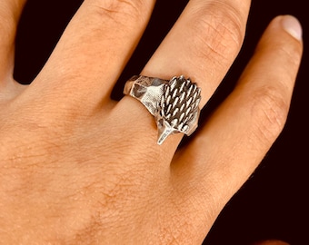 May include: A silver ring featuring a detailed echidna design. The ring showcases the animal's spiky back and pointed snout. The ring has a textured band and is worn on a finger.