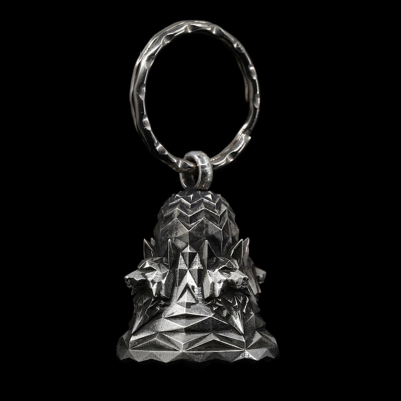 May include: A silver key chain with a bell-shaped charm featuring a geometric design and three wolf heads.