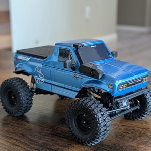 Op de afbeelding: Een blauwe Redcat Ascent RC-truck met grote zwarte offroad-banden. De modeltruck heeft een zwarte rolbeugel en een zwarte bedekking. De truck staat op een houten oppervlak.
