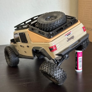 Op de afbeelding: Een beige speelgoedjeep met een zwart dakrek en een reservewiel gemonteerd op de achterkant. De Jeep heeft zwarte wielen en een "Jeep" logo op de achterkant.