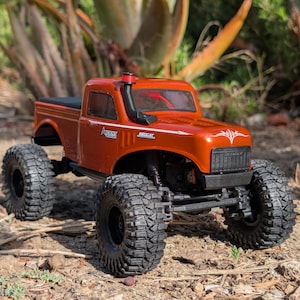 Op de afbeelding: Een oranje RC-truck met grote zwarte banden. De truck heeft een zwarte rolbeugel en een zwarte grille. De teksten "ASCENT" en "REDCAT" zijn zichtbaar op de zijkant. De truck staat op aarde en kleine planten.