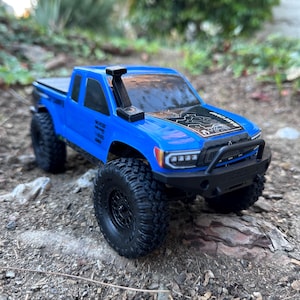 Puede incluir: Un camión de juguete azul con ruedas negras y una jaula de seguridad negra. El camión tiene un gráfico en blanco y negro en el capó y un snorkel negro.