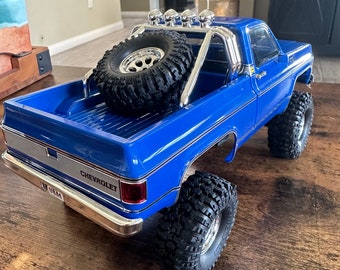 May include: A blue Chevrolet pickup truck with a roll bar and a spare tire in the bed. The truck has a black bumper and black wheels.