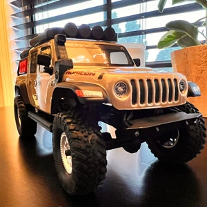 May include: A beige toy Jeep Wrangler with a black roof rack and black wheels. The Jeep has a black front bumper and a black grill with a silver Ruicon logo.