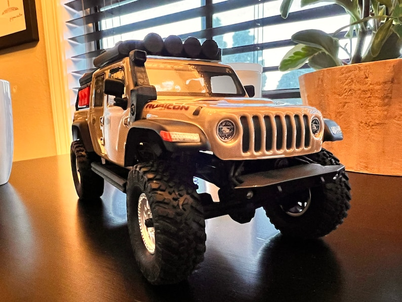 May include: A beige toy Jeep Wrangler with a black roof rack and black wheels. The Jeep has a black front bumper and a black grill with a silver Ruicon logo.
