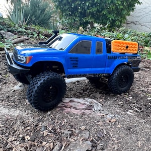 Puede incluir: Un camión teledirigido azul con ruedas y neumáticos negros. El camión tiene un snorkel negro y una caja de almacenamiento naranja. El texto "BASE CAMP" es visible. El camión está sobre una superficie de tierra.