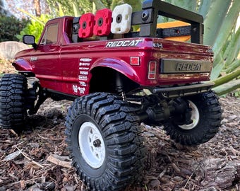 Può includere: Un camioncino rosso e nero con un roll bar nero e una scatola bianca e rossa sul retro. Il camioncino è su un letto di pacciame marrone.