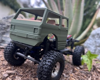 May include: A green model of a van with large, black off-road tires. The van has a detailed body with windows and interior seating. The model is set on a bed of wood chips with greenery in the background.