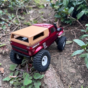Może przedstawiać: Czerwono-beżowy samochód zdalnie sterowany z twardym plastikowym nadwoziem i beżowym dachem. Samochód znajduje się na polnej drodze z zielonymi roślinami w tle. Samochód ma napis "REDCAT" nadrukowany na boku.