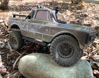 May include: A gray and black toy truck with a black roll bar and a black snorkel. The truck is sitting on a rock with brown mulch in the background.