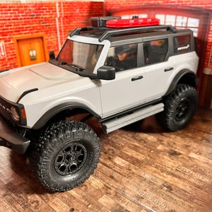 Könnte beinhalten: Ein weißer Ford Bronco mit schwarzen Rädern und einem roten Dachgepäckträger. Das Auto steht auf einem Holzboden.