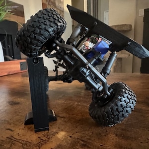 May include: A black and gray toy truck with large tires is displayed on a black stand. The truck is tilted on its side, showing the underside of the chassis. The truck has a black body and a black spoiler.