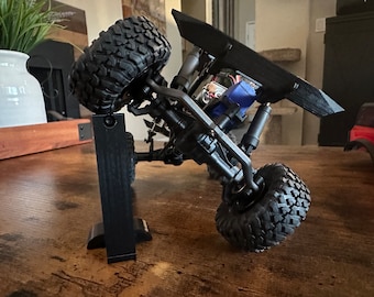 May include: A black and gray toy truck with large tires is displayed on a black stand. The truck is tilted on its side, showing the underside of the chassis. The truck has a black body and a black spoiler.