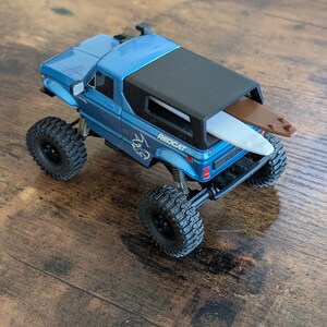 Pode incluir: Um camião de brinquedo Redcat azul com uma cobertura de cama preta e pneus pretos grandes. Uma pequena prancha de surf castanha e branca está presa à parte traseira do camião. O camião está numa superfície de madeira.