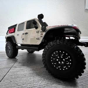 Könnte beinhalten: Ein beiger Jeep Wrangler mit schwarzen Rädern und Reifen, einem schwarzen Überrollkäfig und roten Benzinkanistern auf der Ladefläche. Der Jeep steht auf einem Fliesenboden.