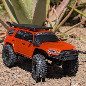 May include: Orange toy SUV with black tires and a roof rack. The front of the vehicle has the word "TOYOTA" printed on it. The vehicle is sitting on a bed of dirt and small rocks.