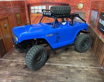 May include: A blue toy truck with a black roll cage and a spare tire on the roof. The truck has black wheels and tires and a KMC sticker on the side.