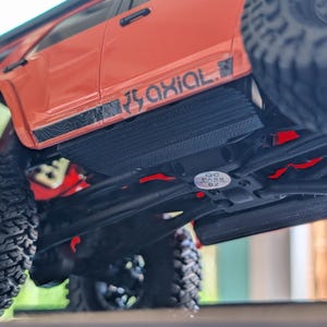 Puede incluir: Vista de cerca de un camión RC Axial rojo y negro. El camión presenta neumáticos todoterreno negros grandes y una parte inferior detallada. El logotipo de Axial es visible en el lateral. Una pegatina QC Pass está presente.