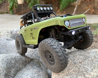 Op de afbeelding: Een groene en zwarte off-road speelgoedtruck met een zwarte rolkooi, een zwarte dakdrager en een zwarte voorbumper. De truck heeft een zwarte grille met het woord "AXIAL" erop gedrukt. De truck staat op een rotsachtig oppervlak.