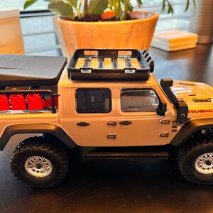 May include: A white Jeep Rubicon pickup truck with a black roof top tent and a black roof rack. The truck has red gas cans on the side and black wheels with silver rims.