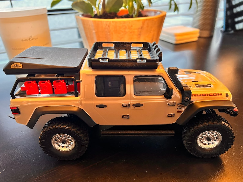 May include: A white Jeep Rubicon pickup truck with a black roof top tent and a black roof rack. The truck has red gas cans on the side and black wheels with silver rims.