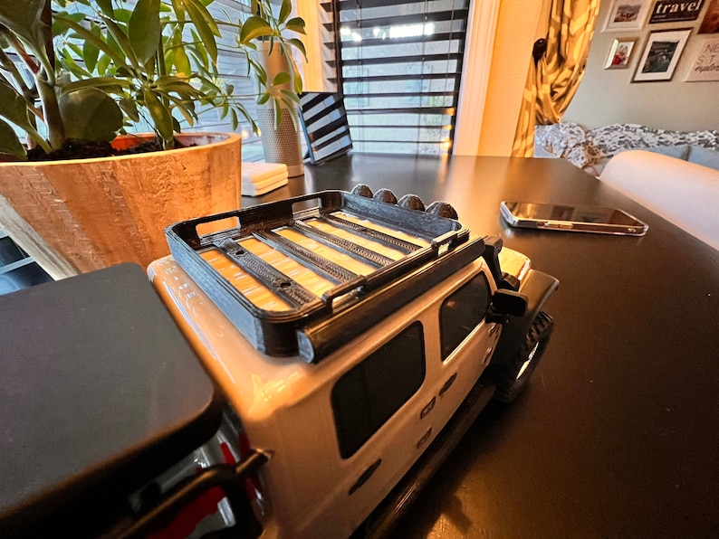 May include: A white toy Jeep with a black roof rack and black wheels. The Jeep is sitting on a dark brown table.