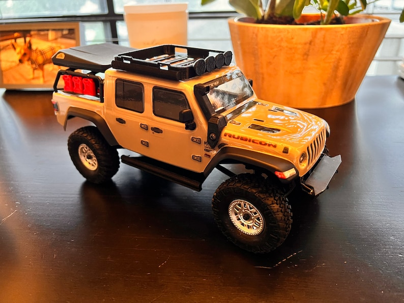 May include: A white Jeep Rubicon toy truck with a black roof rack and red gas cans. The truck has black wheels and a black bumper. The truck is sitting on a dark brown surface.