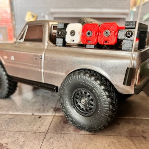 Peut inclure: Un camion pick-up argenté avec un porte-bagages noir et trois bidons de carburant. Les bidons sont rouges, blancs et noirs. Le camion a des roues et des pneus noirs.