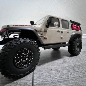 Könnte beinhalten: Ein beiger Jeep Rubicon Pickup mit schwarzen Rädern und Reifen. Der Wagen hat ein schwarzes Dachgepäckträger mit einem roten Benzinkanister oben drauf. Der Wagen steht auf einem hellen Holzboden.