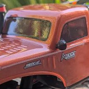 Op de afbeelding: Een metallic oranje op afstand bestuurbare vrachtwagencarrosserie met een zwarte snorkel op de motorkap, een zwarte zijspiegel en stickers met de tekst Redcat en Ascent. Het voertuig heeft een glanzende afwerking met witte strepen.