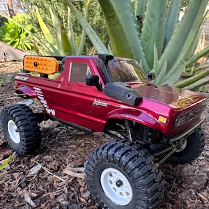 Puede incluir: Un camión de juguete rojo con detalles negros y ruedas blancas. El camión tiene una jaula de seguridad negra y una caja negra con un accesorio naranja y negro montado en la parte trasera. El camión está sentado en un lecho de astillas de madera marrones.