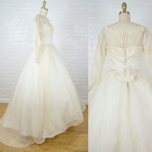 May include: A vintage ivory wedding gown with long sleeves and a full skirt. The bodice features a delicate lace neckline and a satin ribbon at the waist. The back has a row of buttons and a floral accent.