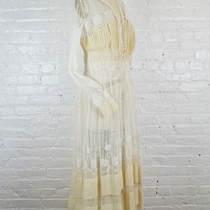 May include: Cream-colored vintage dress with a sheer overlay and intricate lace detailing. The dress features a high neckline, long sleeves, and a tiered skirt. The bodice is fitted, and the dress is displayed on a mannequin against a white brick wall.