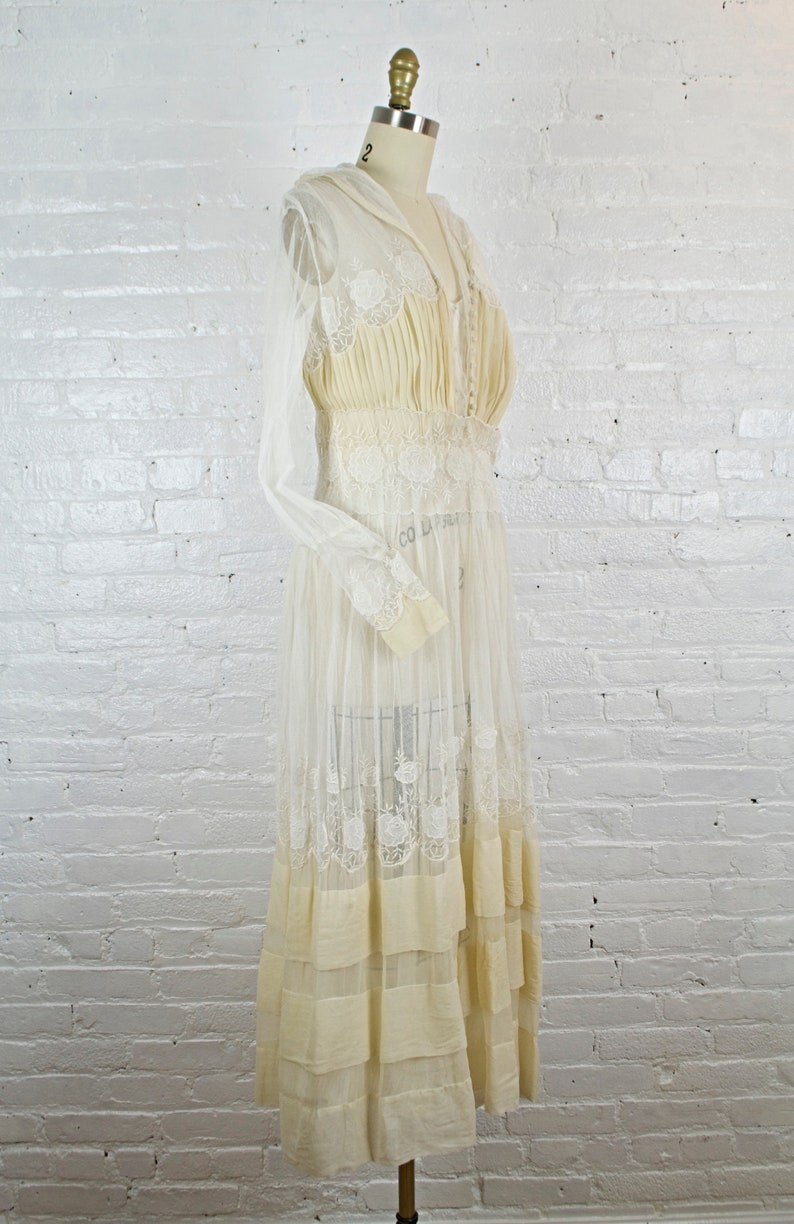 May include: Cream-colored vintage dress with a sheer overlay and intricate lace detailing. The dress features a high neckline, long sleeves, and a tiered skirt. The bodice is fitted, and the dress is displayed on a mannequin against a white brick wall.
