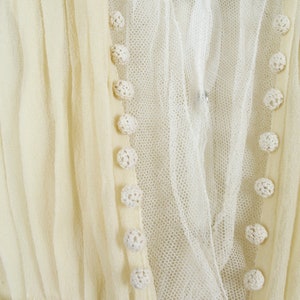 May include: Close-up of a cream-colored vintage dress. The dress features delicate crochet buttons, a sheer overlay, and pleated fabric. The neckline is adorned with lace, and the overall design suggests a romantic, antique aesthetic.