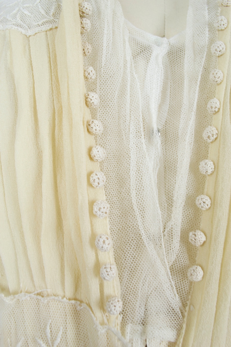 May include: Close-up of a cream-colored vintage dress. The dress features delicate crochet buttons, a sheer overlay, and pleated fabric. The neckline is adorned with lace, and the overall design suggests a romantic, antique aesthetic.