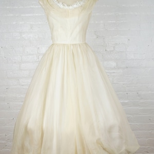 May include: Ivory vintage dress on a mannequin. The dress features a scoop neckline with delicate lace trim, a fitted bodice, and a full, flowing skirt. The skirt is made of a sheer, layered fabric that creates a voluminous silhouette. The dress is displayed against a white brick wall.